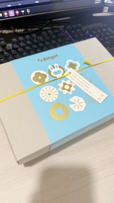 A Bitget-branded gift box wrapped with a yellow ribbon and decorated with cartoon stickers, sitting on a desk in front of a keyboard.