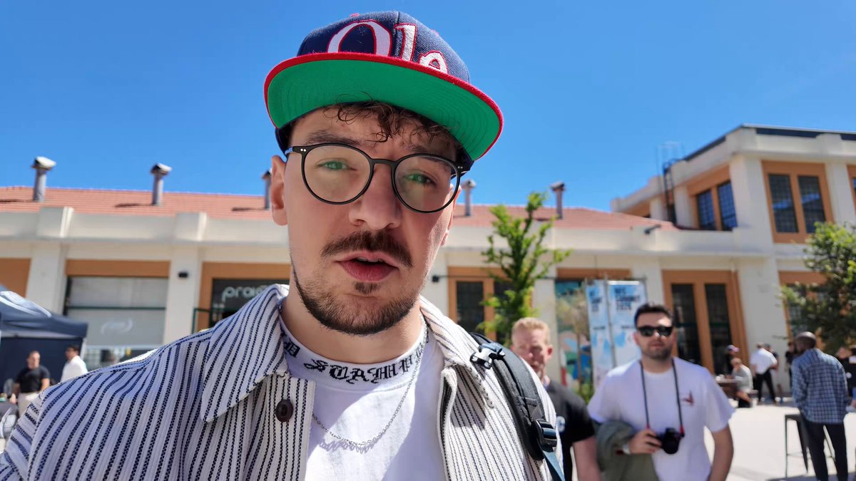 The image shows a man wearing glasses, a striped shirt, and a baseball cap with the word "Ole" written on it. He has his mouth open as if he's speaking or making a face for the camera. The background features an orange building, which is likely a landmark in Lisbon, Portugal. There are other people present in the scene, but they are not the main focus of the image.