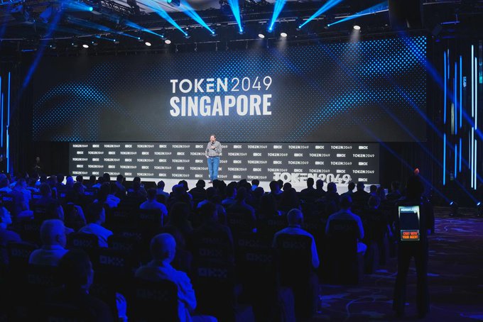 A speaker presents on stage at the TOKEN2049 SINGAPORE conference, with a large illuminated backdrop displaying the event name and seated audience members visible in the foreground.