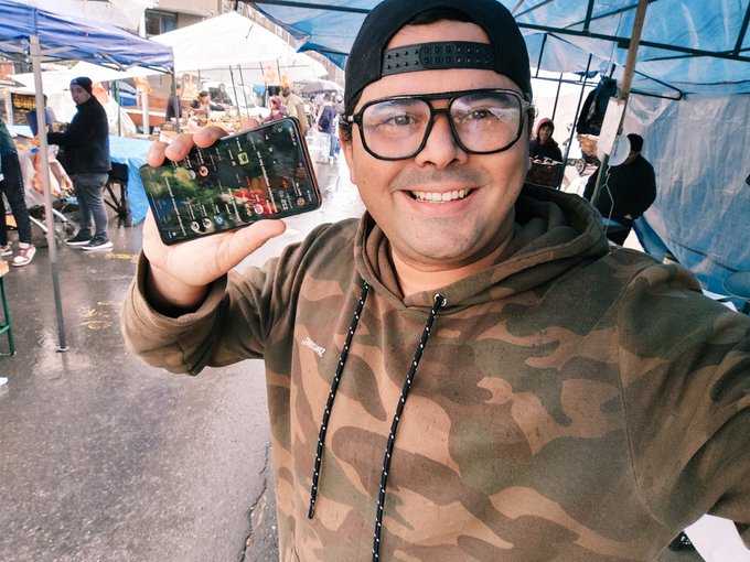 A man wearing a camouflage hoodie, glasses, and a backwards cap smiles while holding up a smartphone displaying the OpenSea app at an outdoor street market in Chile, where tents and other shoppers can be seen in the background despite the rainy 8°C weather.
