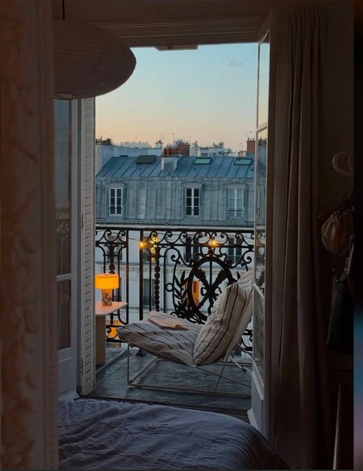 A view from inside a bedroom looking out onto a Parisian balcony with a chair, string lights, and classic rooftop buildings at dusk.