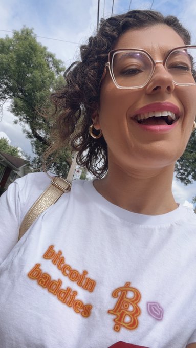 A selfie of a smiling woman with curly hair and glasses wearing a white t-shirt that reads 'bitcoin baddies' with Bitcoin and Ethereum logos.