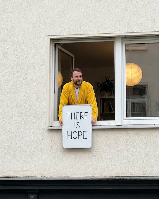 A man in a yellow cardigan leans out an apartment window holding a sign that reads "THERE IS HOPE," a heartfelt real-world message of encouragement that mirrors the post's break from digital screens to share optimism in person.