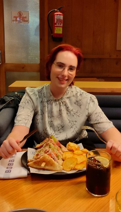 A smiling woman with red hair and glasses sits at a wooden table in front of a club sandwich, chips, and a glass of cola, with a fire extinguisher visible on the wall behind her.
