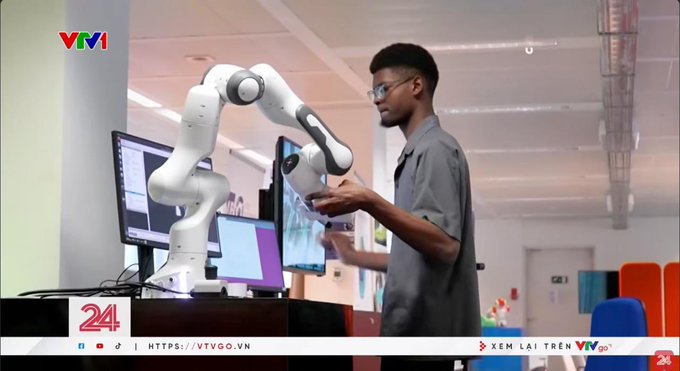 A VTV1 news broadcast screenshot showing a young man wearing glasses and a gray shirt interacting with a white robotic arm (labeled FRANKA) at a desk with computer monitors. The broadcast includes a '24' logo and URL HTTPS://VTVGO.VN.