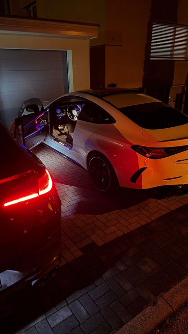 A white BMW coupe parked at night with its driver's door open, illuminated by colorful interior and red exterior lighting on a paved driveway.
