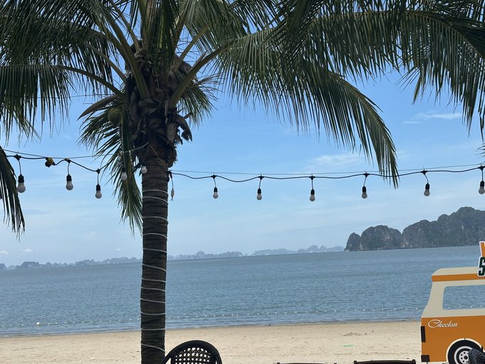 A tropical beach scene featuring a palm tree, string lights, a yellow food truck labeled "Checkin", and limestone karsts rising from the sea in the distance.