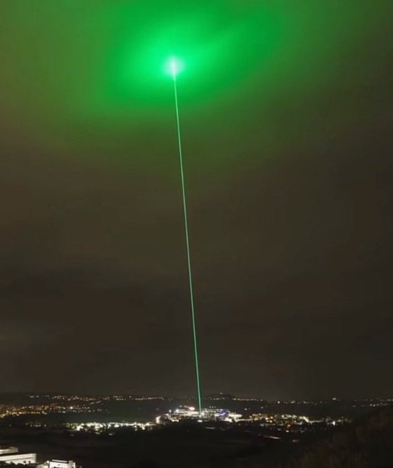 A green laser beam shoots skyward from a brightly lit facility below, creating an aurora-like glow in the night sky above a distant cityscape, visually symbolizing the post's question about which memecoin will skyrocket "to the moon."