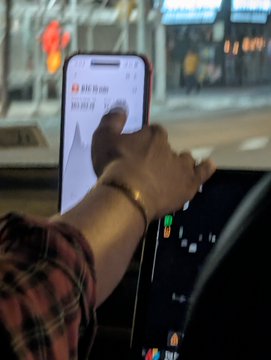 A person's hand, wearing a gold bracelet, reaches from the driver's seat of a car to interact with a large vertical touchscreen infotainment display showing what appears to be an Uber or rideshare driver app with a fare estimate. The scene is captured at night with city lights visible through the windshield.