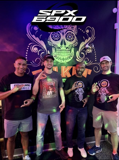 Four men posing in front of an SPX 6900 banner featuring a green sugar skull design, all making hand gestures under purple and green event lighting.