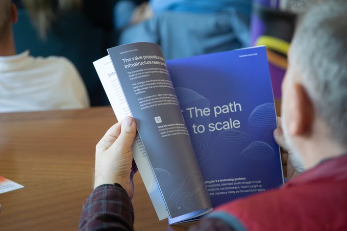 An over-the-shoulder view of a person reading a brochure titled 'The path to scale' with the iQI logo and the headline 'The value proposition infrastructure needs to...'
