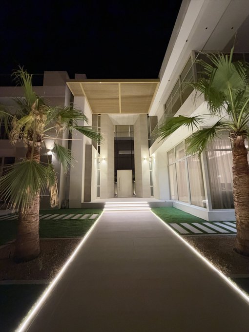 A nighttime view of a modern luxury home exterior featuring illuminated walkways, palm trees, and contemporary architecture.