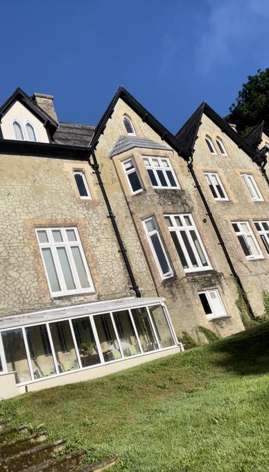 A photo of a large stone manor or estate house with multiple gables, tall windows, and a sunroom extension at the base, set on a grassy lawn under a blue sky.