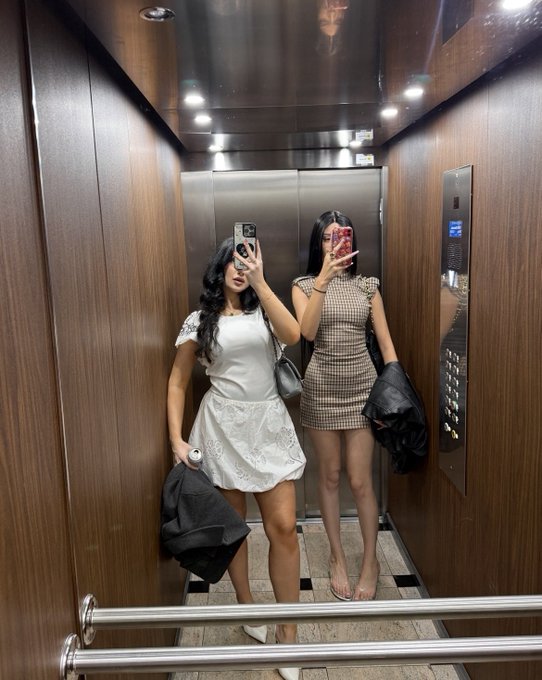 Two young women take mirror selfies inside a wood-paneled elevator; the woman on the left wears a white top and skirt while the woman on the right wears a checkered houndstooth dress.
