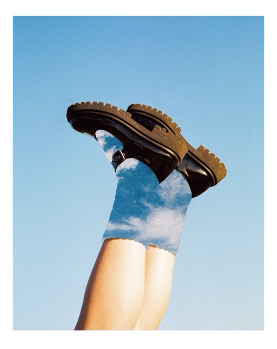 A creative photograph showing legs wearing chunky brown boots pointing up against a blue sky, with a cloud-patterned background.