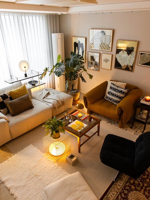 A cozy living room with a beige sofa, brown leather armchair, gallery wall of framed art, a coffee table with snacks, and warm lamp lighting.