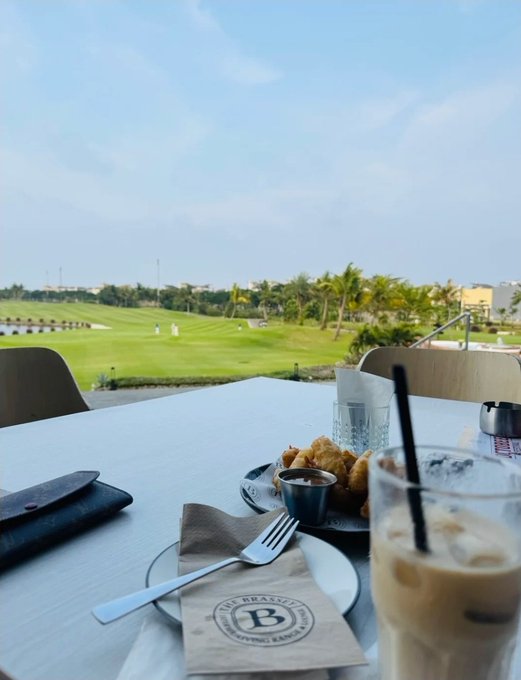 A dining table overlooks a green golf course, set with a plate of food, a coffee glass, cutlery, and a napkin bearing a 'B' logo.