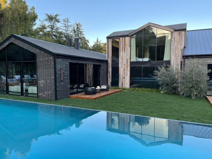 A modern architectural home featuring dark brick, wood paneling, large glass windows, and a reflective swimming pool in the foreground.