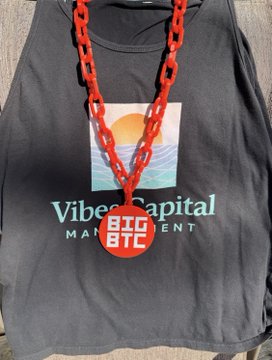 A black "Vibe Capital Management" t-shirt with a sun-over-waves graphic is displayed beneath a chunky orange chain necklace featuring a round red "BIG BTC" medallion, visually reinforcing the bold "Big Bitcoin. Expect us." slogan with bold, statement-making crypto flair.