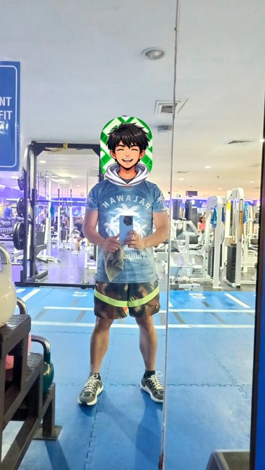 A mirror selfie of a person in a gym wearing a Hawaiian-themed t-shirt and shorts, with an anime-style face filter featuring black hair and cat ears applied over their head.