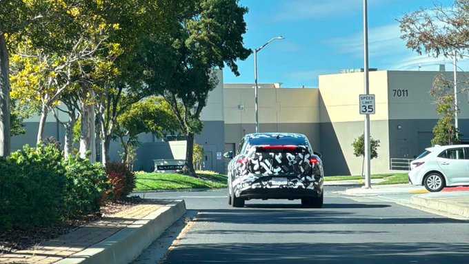 A camouflaged SUV drives down a street with a 35 mph speed limit sign, heading toward a building marked '7011' on a sunny day with clear blue skies.