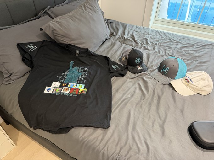 A black t-shirt with a New York City Statue of Liberty graphic and three baseball caps (two New York Yankees caps and one white cap) are arranged on a gray bed.