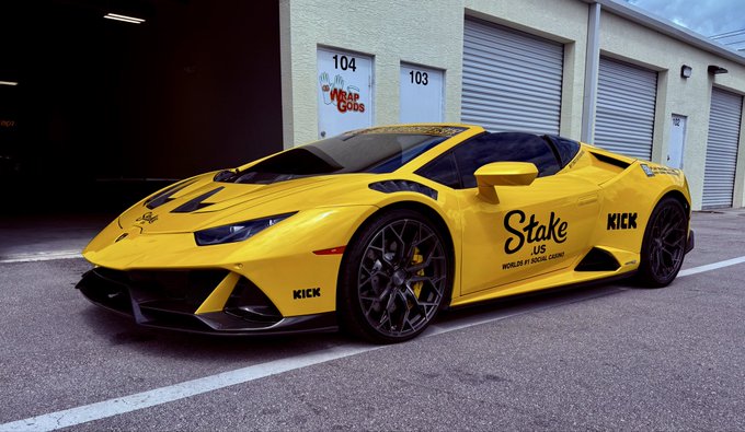 A yellow Lamborghini Huracán wrapped with "Stake.us" and "Kick" branding sits parked in front of garage bays numbered 103 and 104, with a "Wrap Gods" sign visible. The image relates to the post by showcasing the Stake.us branding, tying into the streamer's promotion of their $5,000 bonus hunt, slot spins, and crypto tipping request on Kick.com.