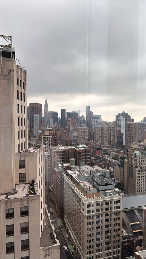 A view of Manhattan's skyline from a high-rise window, with the Empire State Building visible in the distance, framed by neighboring buildings under a cloudy sky. The image doesn't directly relate to the post about Coinbase ($COIN) launching on Base, but it may symbolize a high-level, strategic vantage point on the crypto exchange's next move.