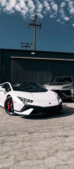 A white Lamborghini is parked on a concrete lot in front of a metal building, with a gray Lamborghini SUV partially visible behind it under a partly cloudy sky.