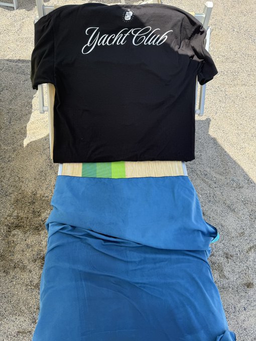 A black t-shirt with 'Yacht Club' printed in white cursive lettering is draped over a chair, with a blue garment or sheet placed below it.