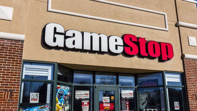 The storefront of a GameStop retail location displays the company's red and white logo prominently above the entrance, with promotional posters advertising "BUY 2 GET 1 FREE" deals visible in the windows. This image relates to the post by connecting to the original GameStop ($GME) phenomenon, which the post references as the catalyst that inspired a blockchain-based utility token built on Binance Smart Chain (BSC).