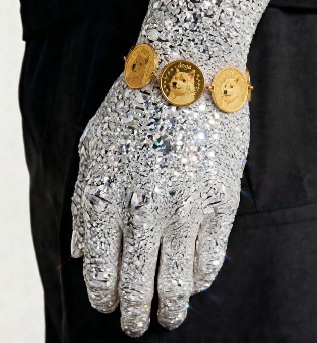 A crystal-encrusted silver glove rests against a dark fabric, adorned with a gold bracelet featuring three Doge coin tokens spelling "DOGE"—a striking visual representation of "diamond hands" in the crypto sense, symbolizing an unbreakable grip on Dogecoin holdings.