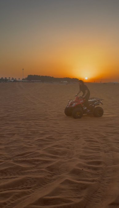A person rides an ATV across a sandy beach at sunset, with footprints visible in the sand.