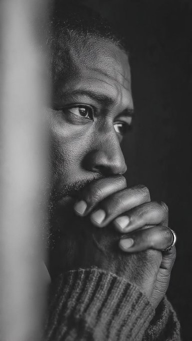 A black and white close-up portrait of a Black man in a sweater holding his hand near his mouth in a contemplative pose.