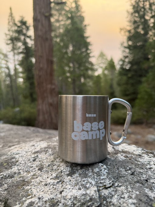 A stainless steel camping mug with "base camp" printed on it sits on a large rock, with tall pine trees and a sunset-lit sky in the background, fitting the poster's message of being away in the mountains for a camping weekend.