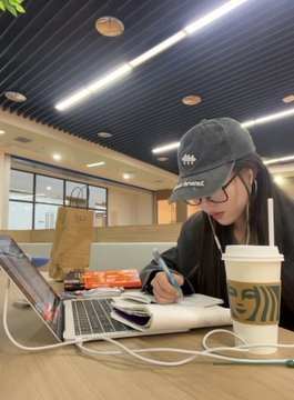 A young woman wearing a gray cap and glasses studies at a wooden table with an open notebook, laptop, and iced coffee, charging her phone via a white cable.