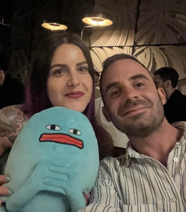 A selfie of a man and woman posing together at what appears to be a restaurant, with the woman holding a blue plush toy.