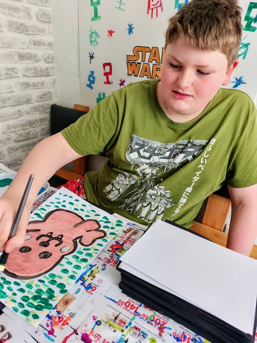 A boy in a green t-shirt sits at a table, focused on painting a pink creature with green spots, with a 'Star Wars' decoration visible on the wall behind him.