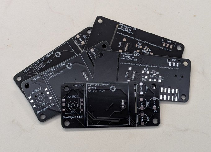 A pile of unassembled black PCBs labeled "SeedSigner 1.54" Design by @NurdRBotCoin" featuring components for a 1.54" LCD 240x240 display with joystick controls, representing the first production batch of the SeedSigner Improvement Proposal 154 (SIP154) hardware upgrade.