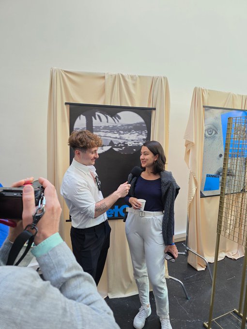 A man in a white shirt and tie is interviewing a woman in a navy top and light pants at an event, with artwork visible in the background.