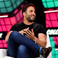 A man with short dark hair and a beard, wearing a black t-shirt and blue jeans, sits cross-legged on a stage while speaking into a headset microphone.