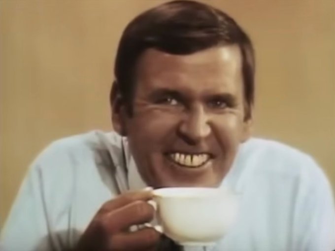 A vintage television-style close-up shows a smiling man in a light-colored shirt holding a white cup, looking directly at the camera with an enthusiastic expression.