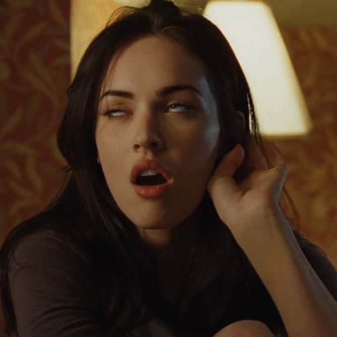 A woman with dark hair and an open-mouthed expression looks to the side with her hand near her face, illuminated by warm light.