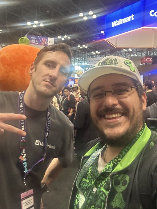 Two smiling men posing at a convention or expo (with 'Walmart' visible overhead), one wearing a black 'Uber' shirt and a lanyard, the other in a green patterned jacket and a white hat with green alien logos.