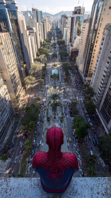 Spider-Man in his classic red and blue suit sits on a ledge looking down over a busy multi-lane city avenue with skyscrapers on either side.