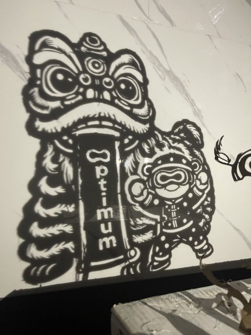 A black and white stencil-style street art mural on a white tarp featuring a traditional Chinese lion dance head with an "Optimum" banner in its mouth, alongside a small character in a spacesuit.