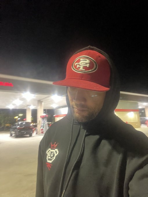 A nighttime selfie of a man wearing a red San Francisco 49ers cap and a dark hoodie, standing at a brightly lit gas station.