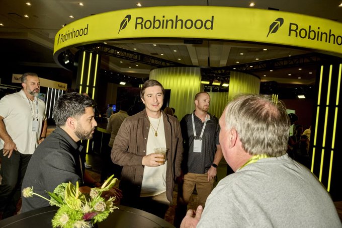 Men converse at a Robinhood booth, identifiable by the prominent yellow signage and feather logo wrapping around the stand.