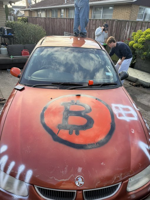 A red Vauxhall car is being spray-painted with a large Bitcoin logo on its hood in a residential driveway, tying into the "$BITCOIN" caption by visualizing cryptocurrency branding. One person stands on the roof while others work around the vehicle, suggesting a creative project celebrating Bitcoin.
