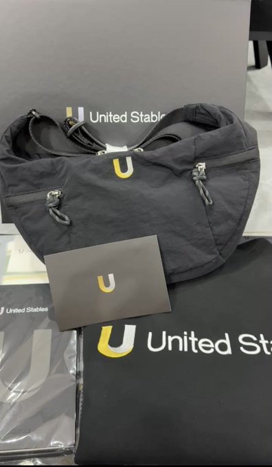 A black "United Stables" branded shoulder bag with a yellow "U" logo, displayed alongside matching merchandise packaging and a card featuring the same logo.

The image showcases the physical merchandise mentioned in the post—specifically the practical bag the user received from the $U (United Stables) stablecoin project community, representing their appreciation for the team's branded swag.